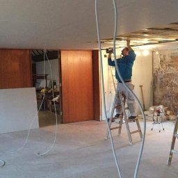 Home & Trade Tomasz Gliński - Pracownik na drabinie montuje płytę karton-gips na suficie w pomieszczeniu z widocznymi przewodami elektrycznymi i elementami wykończeniowymi.