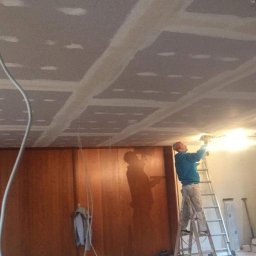 Home & Trade Tomasz Gliński - Pracownik na drabinie szpachluje łączenia płyt karton-gips na suficie. Widoczne narzędzia, wiadro i przewody. Ściana z boazerią w tle.