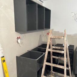 Home & Trade Tomasz Gliński - Częściowo zamontowane, grafitowe szafki kuchenne wiszące i stojące, drabina malarska, poziomica Stanley i wystające kable elektryczne na tle surowej ściany.