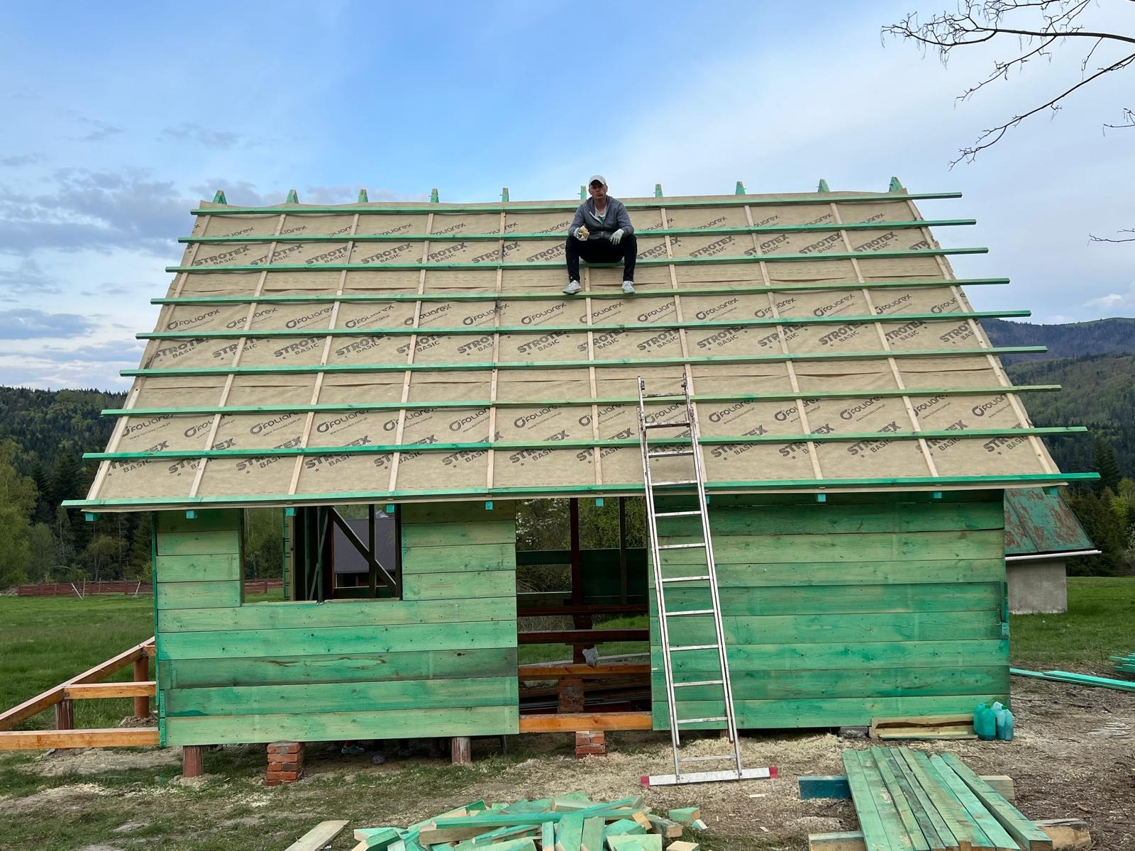 Budowa szkieletu domu z zielonych desek i folii Strotex na dachu, drabina oparta o ścianę, osoba siedząca na dachu, widok na góry w tle.