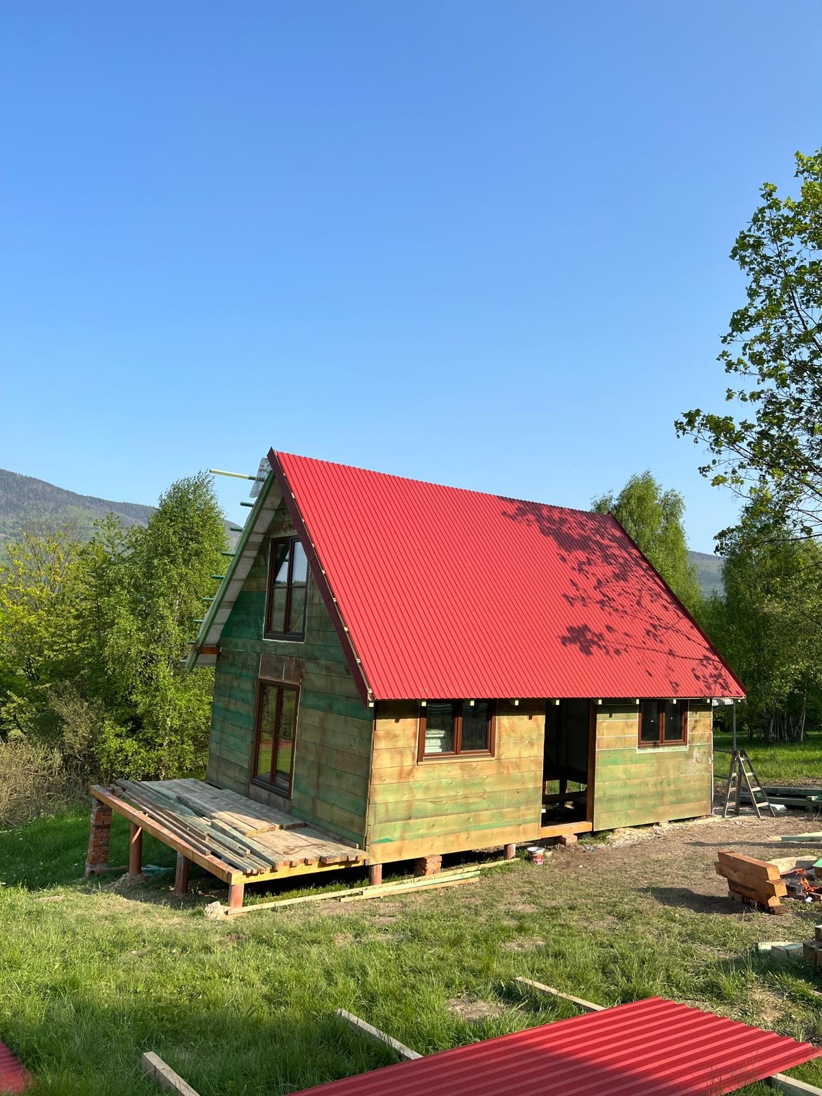 Drewniany dom z czerwonym dachem w trakcie budowy, widok z zewnątrz, w tle zieleń i góry. Częściowo wykonana elewacja i taras. Słoneczny dzień.