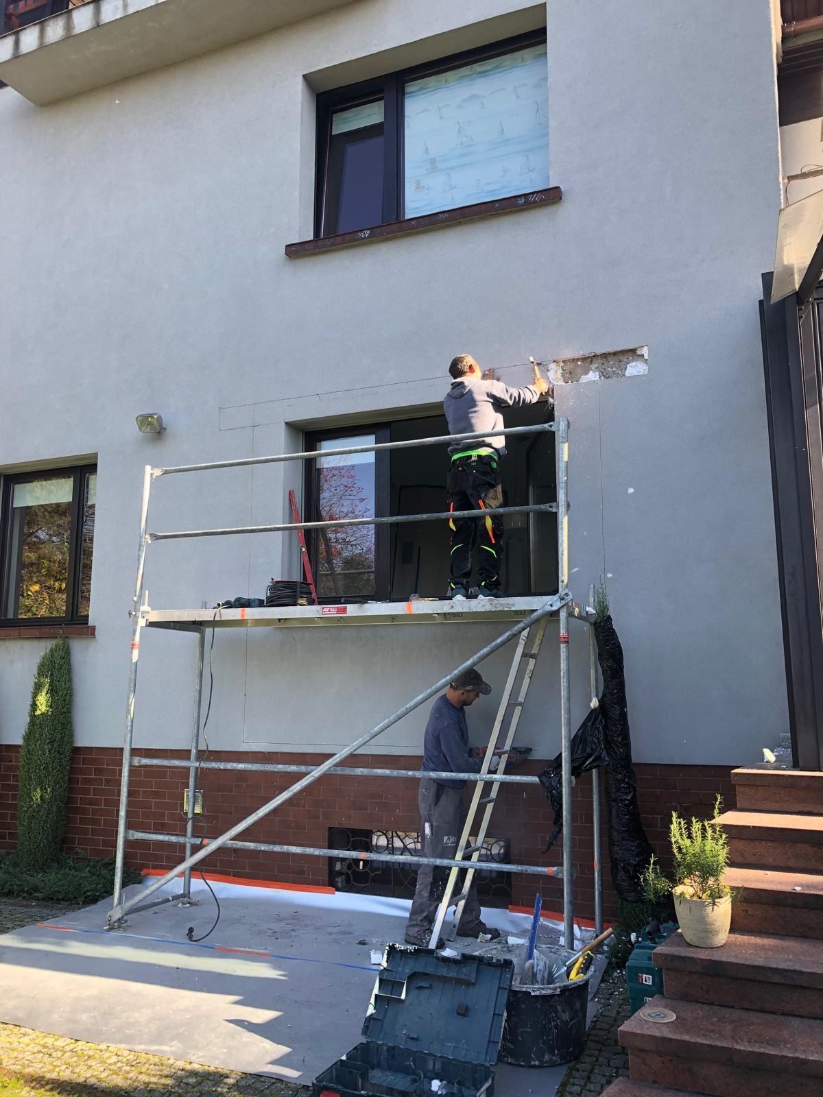 Pracownicy na rusztowaniu przygotowują otwór w elewacji budynku pod montaż okna. Widoczne narzędzia i materiały budowlane. Praca w toku, jasne oświetlenie.