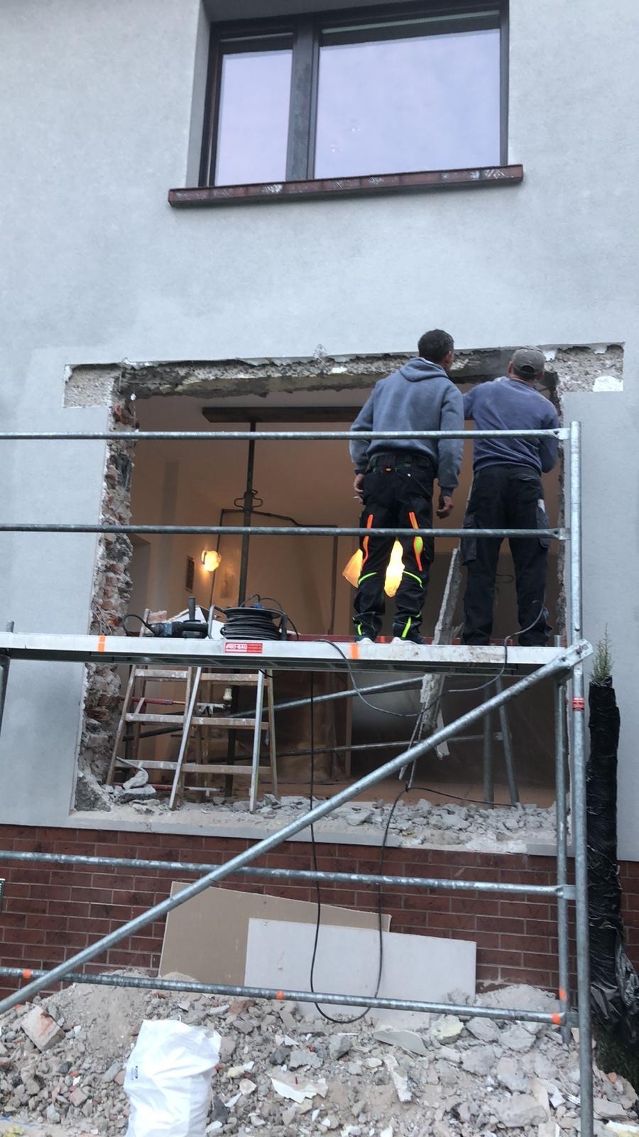 Dwóch pracowników na rusztowaniu wykuwa otwór okienny w szarej elewacji domu. Widoczne narzędzia, gruz i cegły. W tle wnętrze z lampą. Praca w toku.