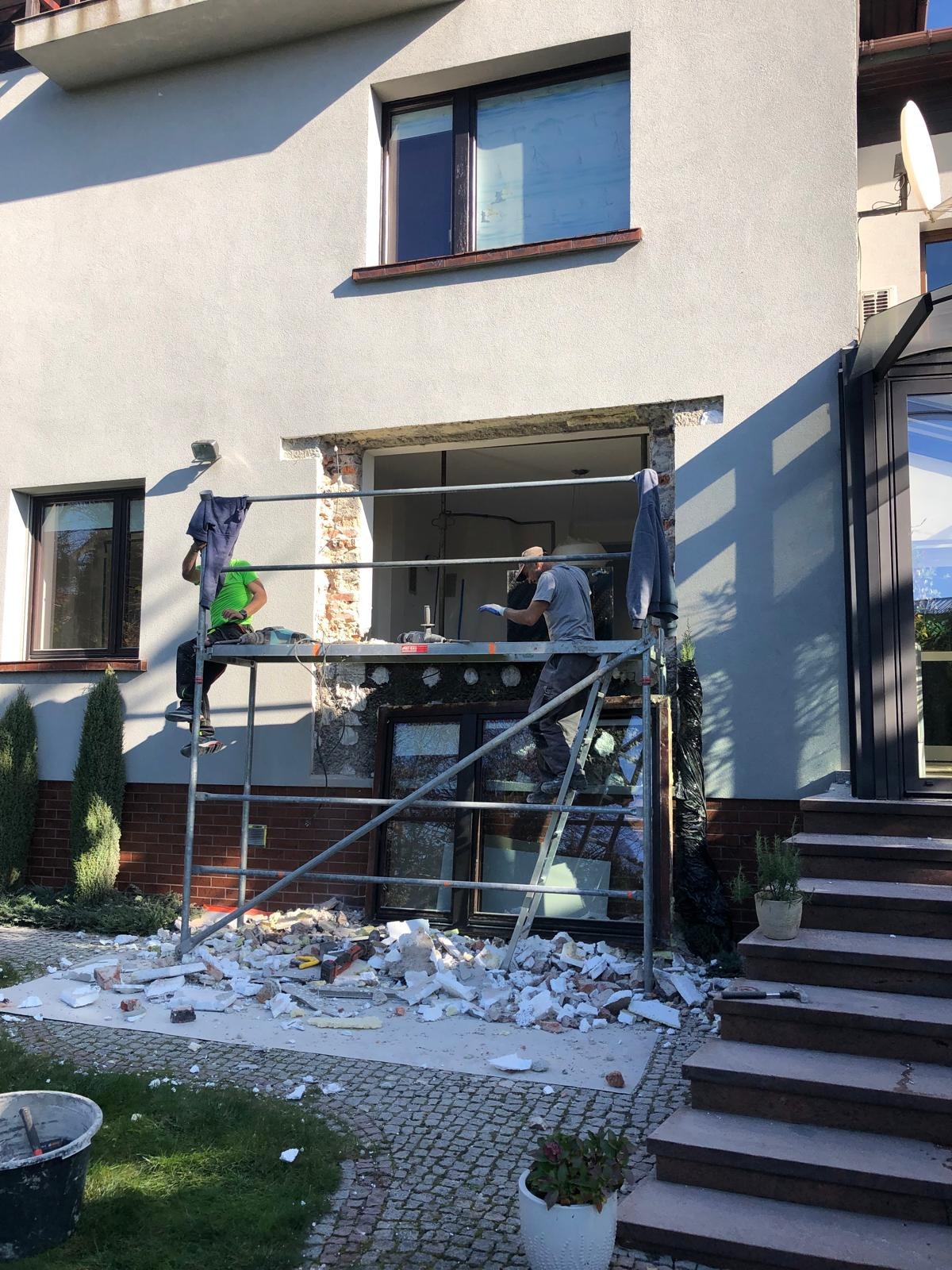 Demontaż okna na rusztowaniu. Dwóch pracowników w trakcie prac przygotowawczych do montażu nowego okna. Gruz i resztki materiałów budowlanych na ziemi.