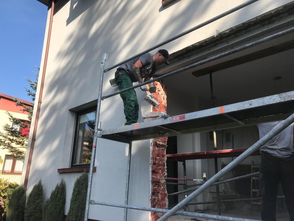Pracownik na rusztowaniu, w trakcie skuwania tynku z elewacji domu jednorodzinnego. Widoczne narzędzia i fragment odsłoniętej cegły. Słoneczny dzień.