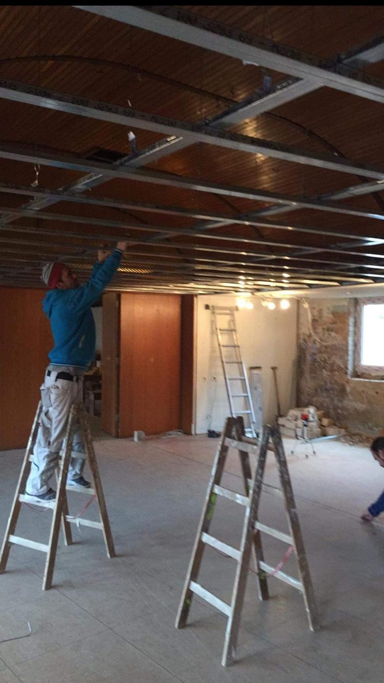 Pracownik na drabinie montuje metalowy stelaż sufitu podwieszanego w remontowanym pomieszczeniu. Widoczne elementy instalacji i surowy stan ścian.