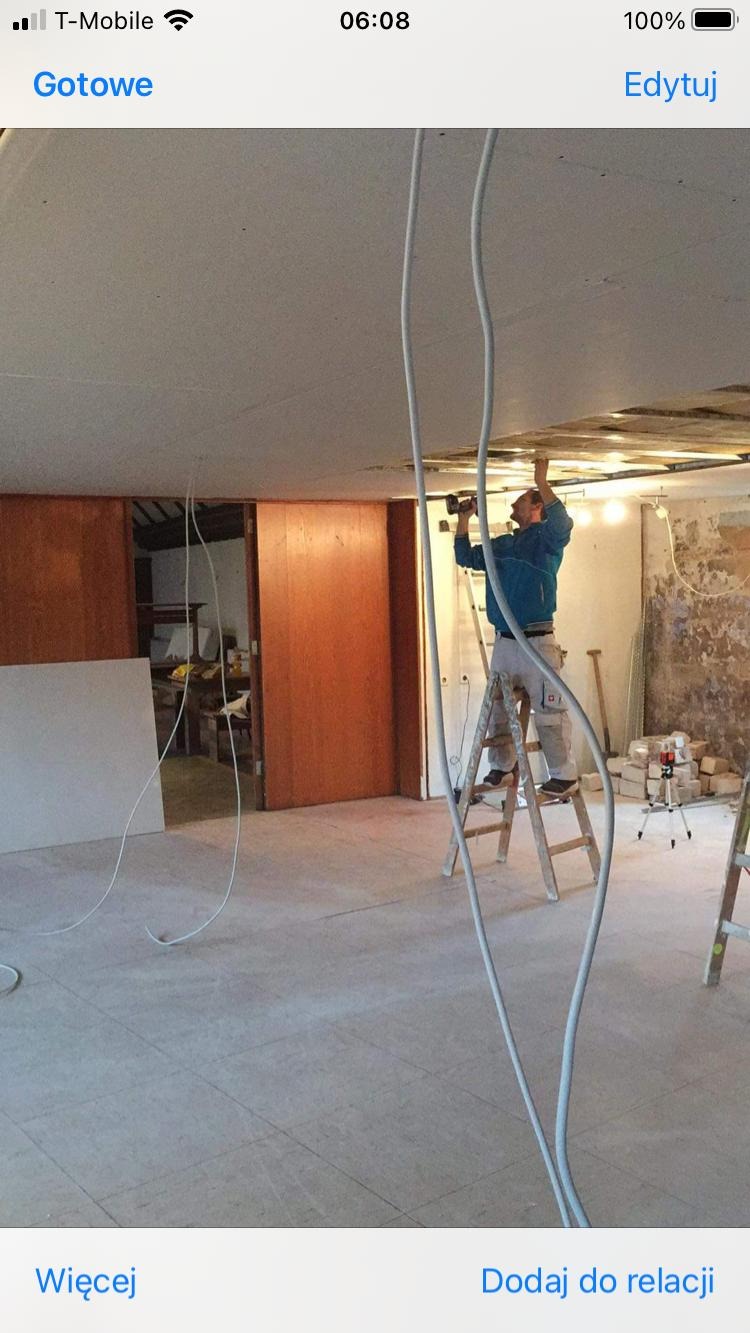 Pracownik na drabinie montuje płytę karton-gips na suficie w pomieszczeniu z widocznymi przewodami elektrycznymi i elementami wykończeniowymi.