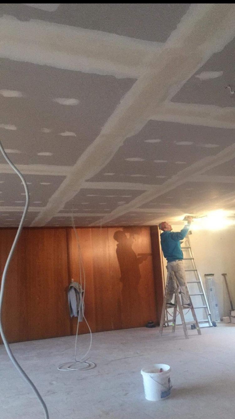 Pracownik na drabinie szpachluje łączenia płyt karton-gips na suficie. Widoczne narzędzia, wiadro i przewody. Ściana z boazerią w tle.