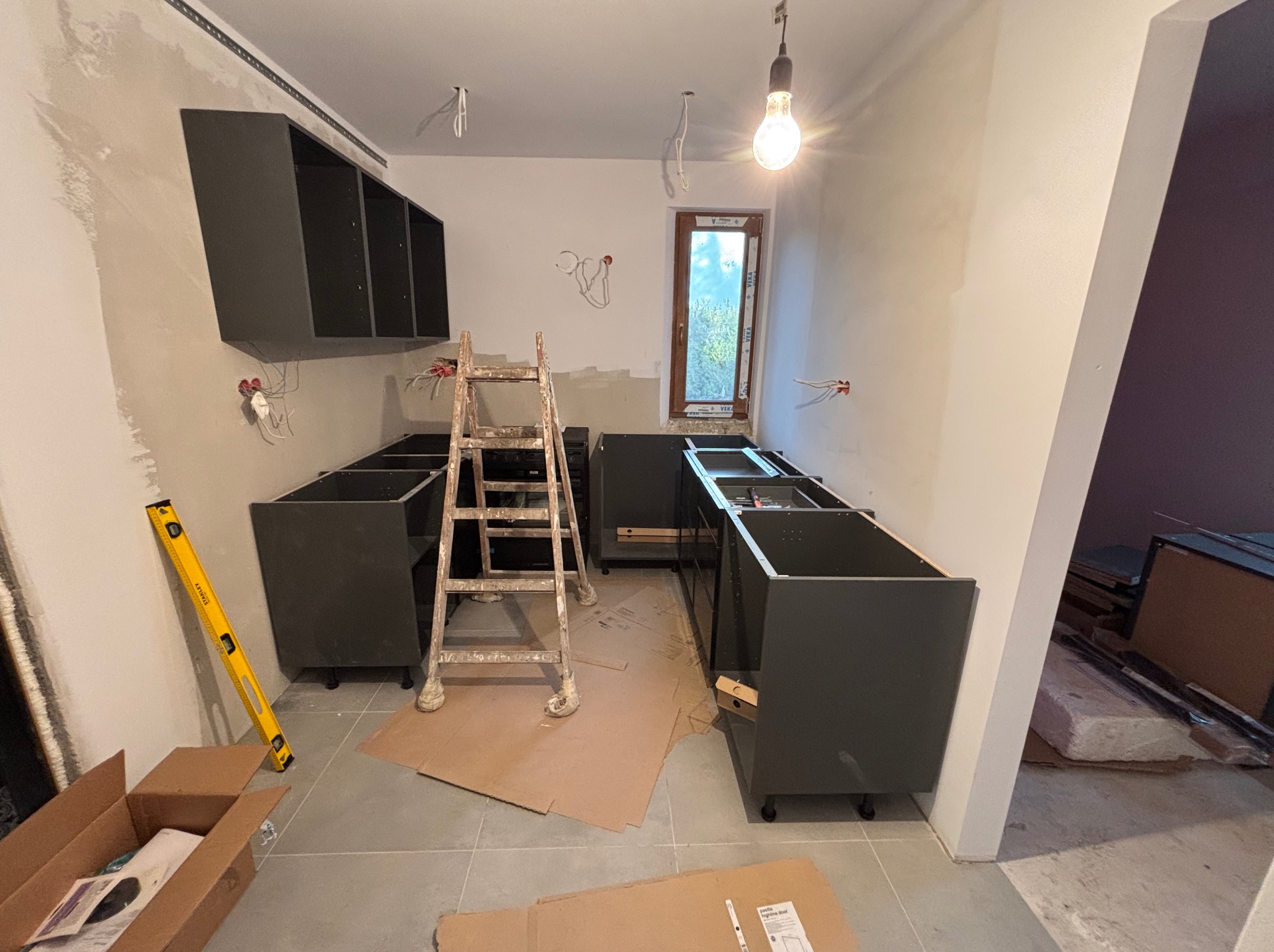 Wnętrze kuchni w trakcie montażu: szare szafki bez blatów, drabina malarska, poziomica przy ścianie, kartony na podłodze. Widoczne instalacje elektryczne.