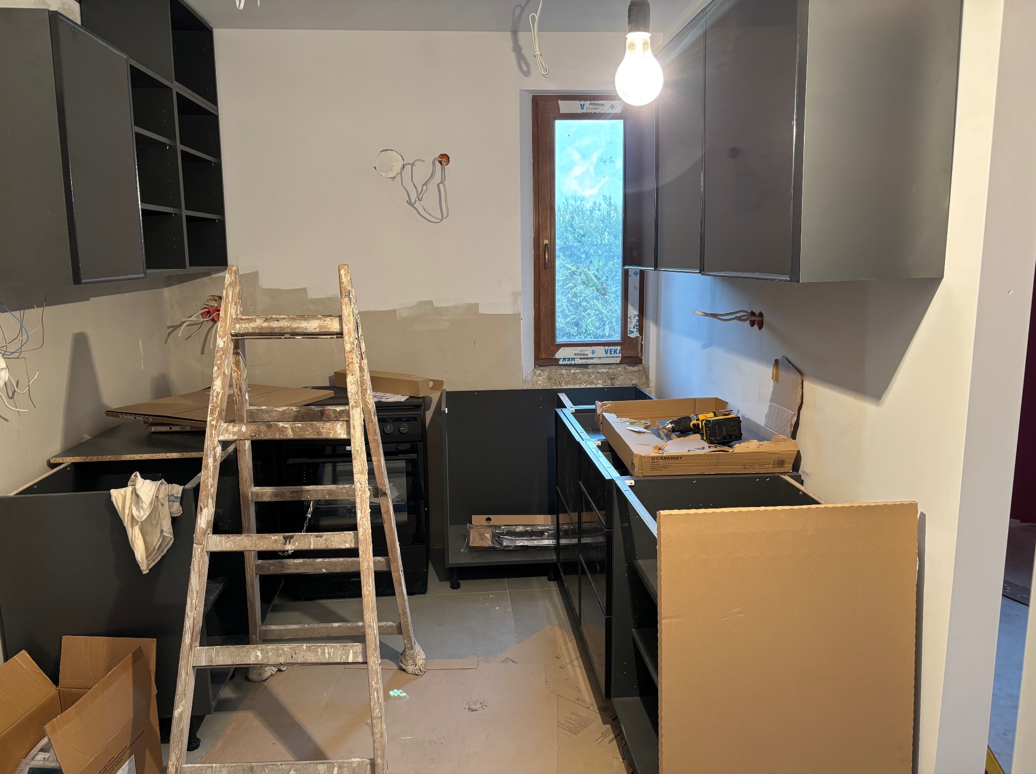 Częściowy montaż czarnych szafek kuchennych w jasnym pomieszczeniu. Widoczna drabina, kartony i narzędzia. W tle okno z widokiem na zieleń.