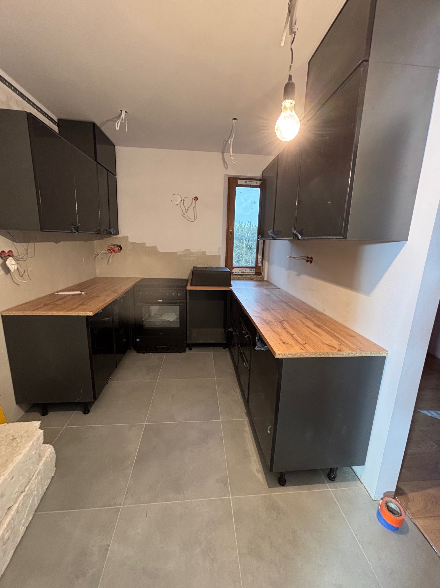 Częściowo zamontowana, czarna zabudowa kuchenna z drewnianym blatem. Widoczne niedokończone ściany i wystające kable. Oświetlenie żarówką. Ujęcie z poziomu podłogi.