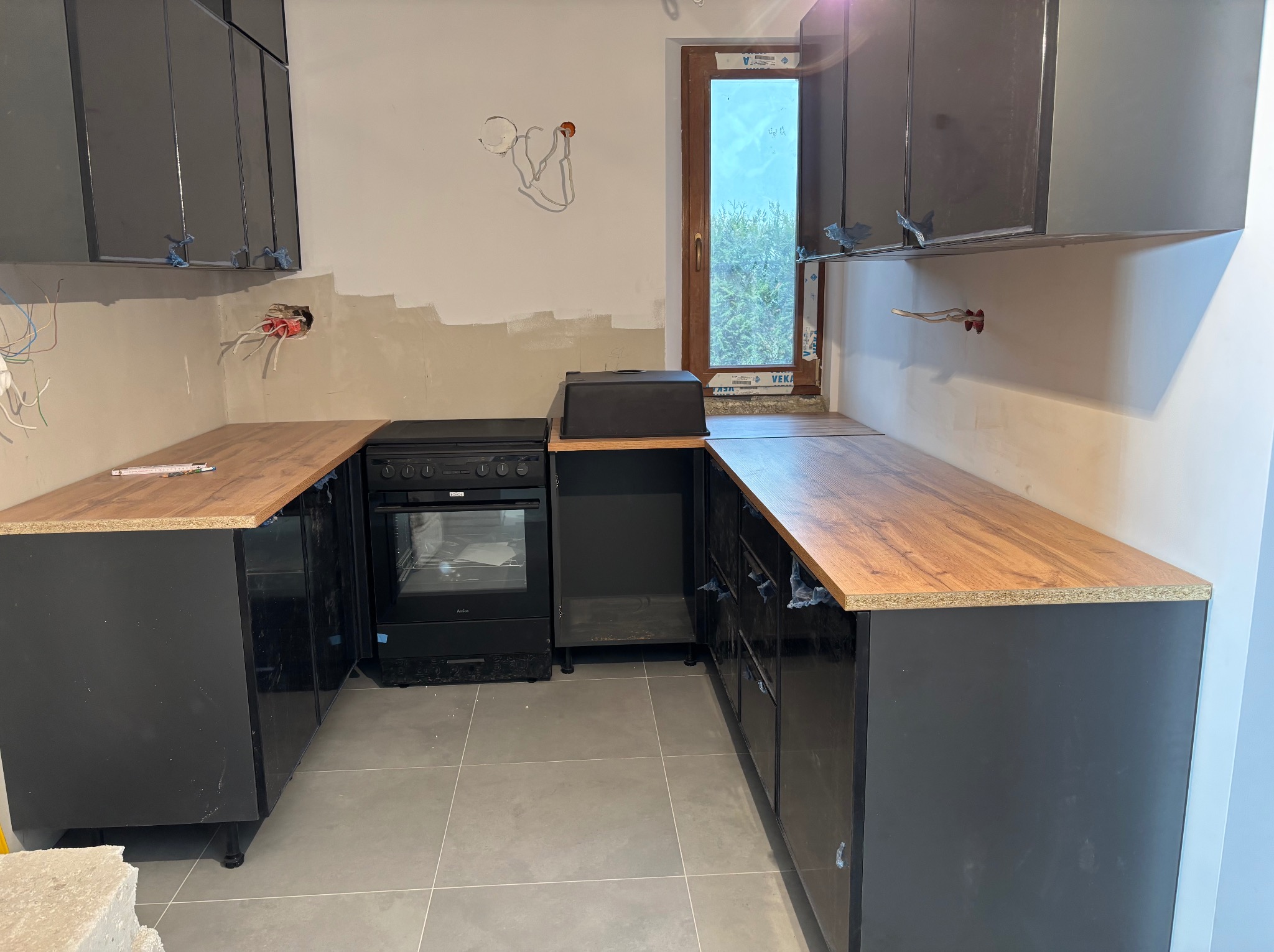Częściowo zamontowana, nowoczesna kuchnia z czarnymi szafkami i drewnianym blatem. Widoczne niedokończone ściany i instalacje elektryczne.