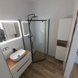 JAN BERGMANN - Nowoczesna łazienka z kabiną prysznicową typu walk-in, umywalką nablatową na drewnianym blacie i podświetlanym lustrem. Białe płytki i drewnopodobna podłoga.