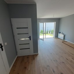 JAN BERGMANN - Jasne drewniane panele podłogowe w szarym pokoju z białymi drzwiami i widokiem na ogród przez otwarte drzwi balkonowe. Minimalistyczny design.