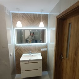 JAN BERGMANN - Nowoczesna łazienka po remoncie: biała szafka z umywalką, podświetlane lustro, drewniana mozaika na ścianie i drewniane drzwi. Widoczne oświetlenie.