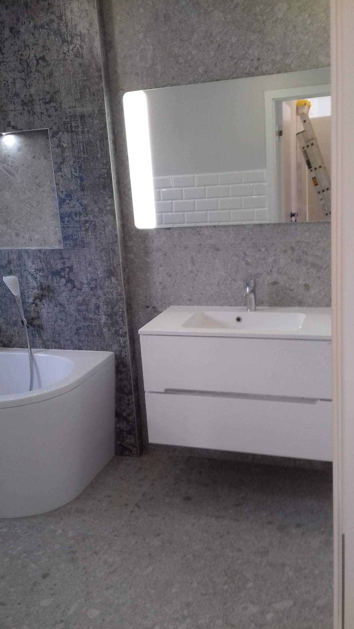 Nowoczesna łazienka po remoncie: wanna z baterią, biała szafka z umywalką i podświetlane lustro. W tle widoczna drabina, co sugeruje świeżo zakończone prace.