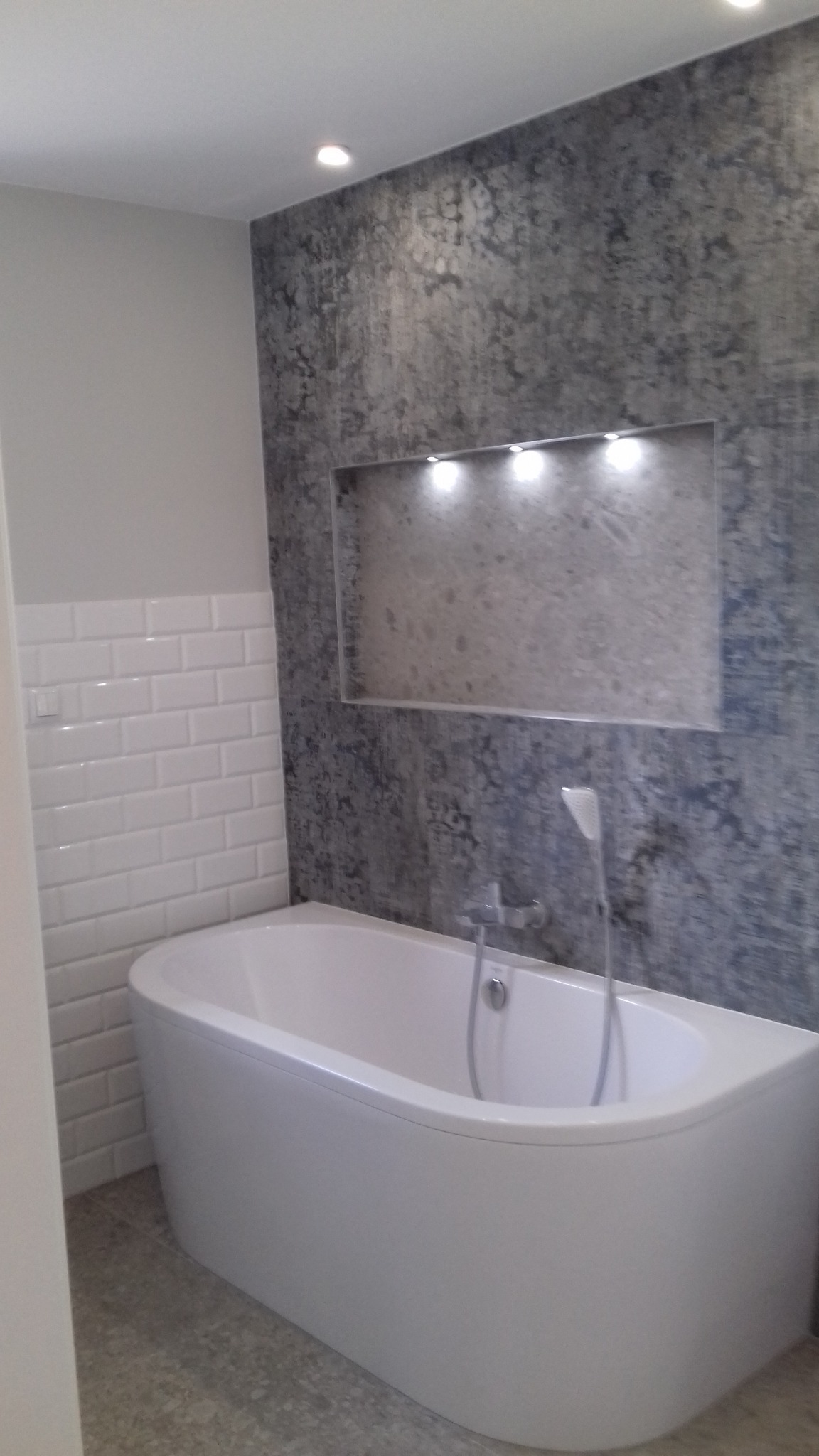 Nowoczesna łazienka z wolnostojącą wanną, białą glazurą typu metro i ścianą z szarego betonu architektonicznego z podświetloną wnęką. Minimalistyczny design.