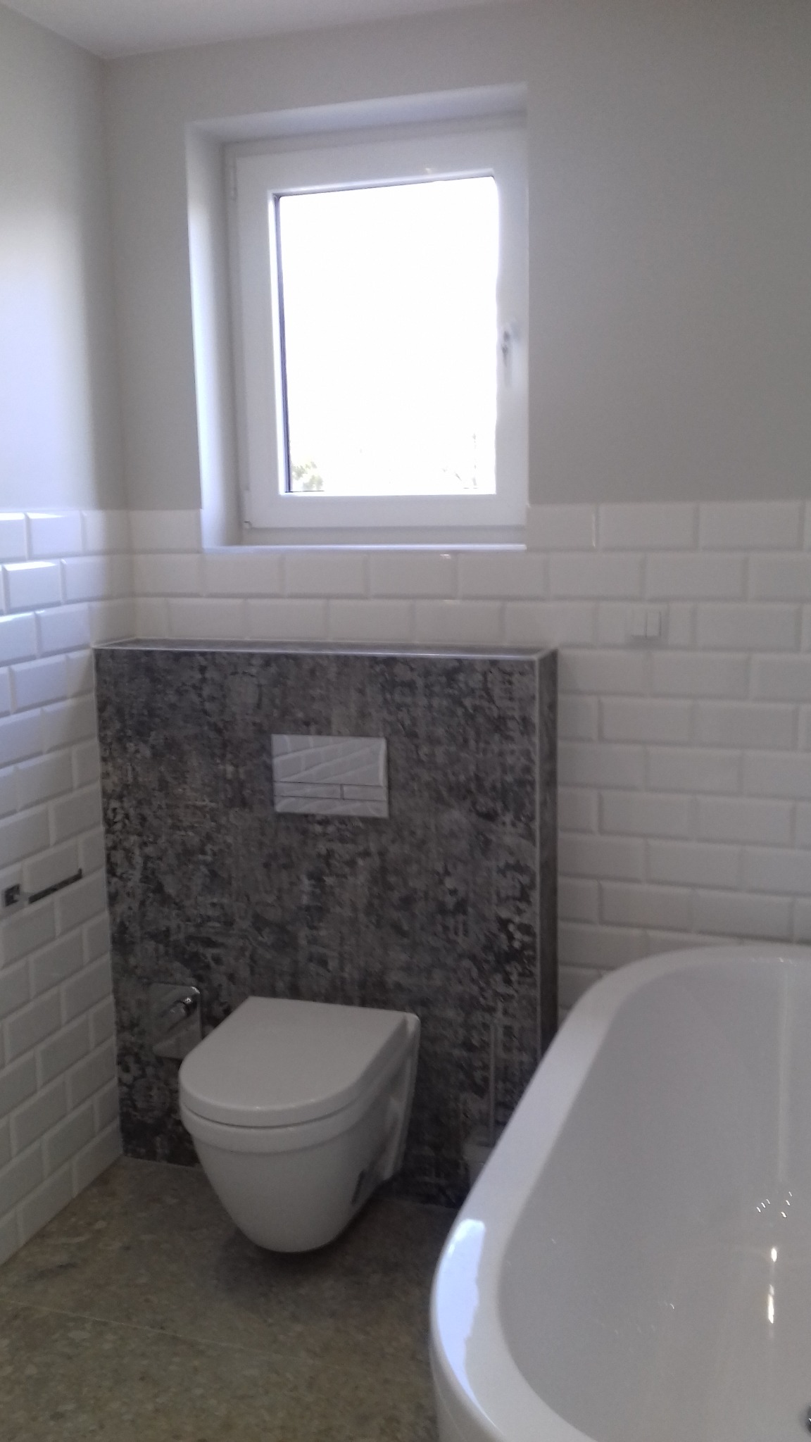 Nowoczesna łazienka z podwieszaną toaletą i wanną, wykończona białymi płytkami typu metro i szarą okładziną za toaletą. Widoczne okno z naturalnym światłem.
