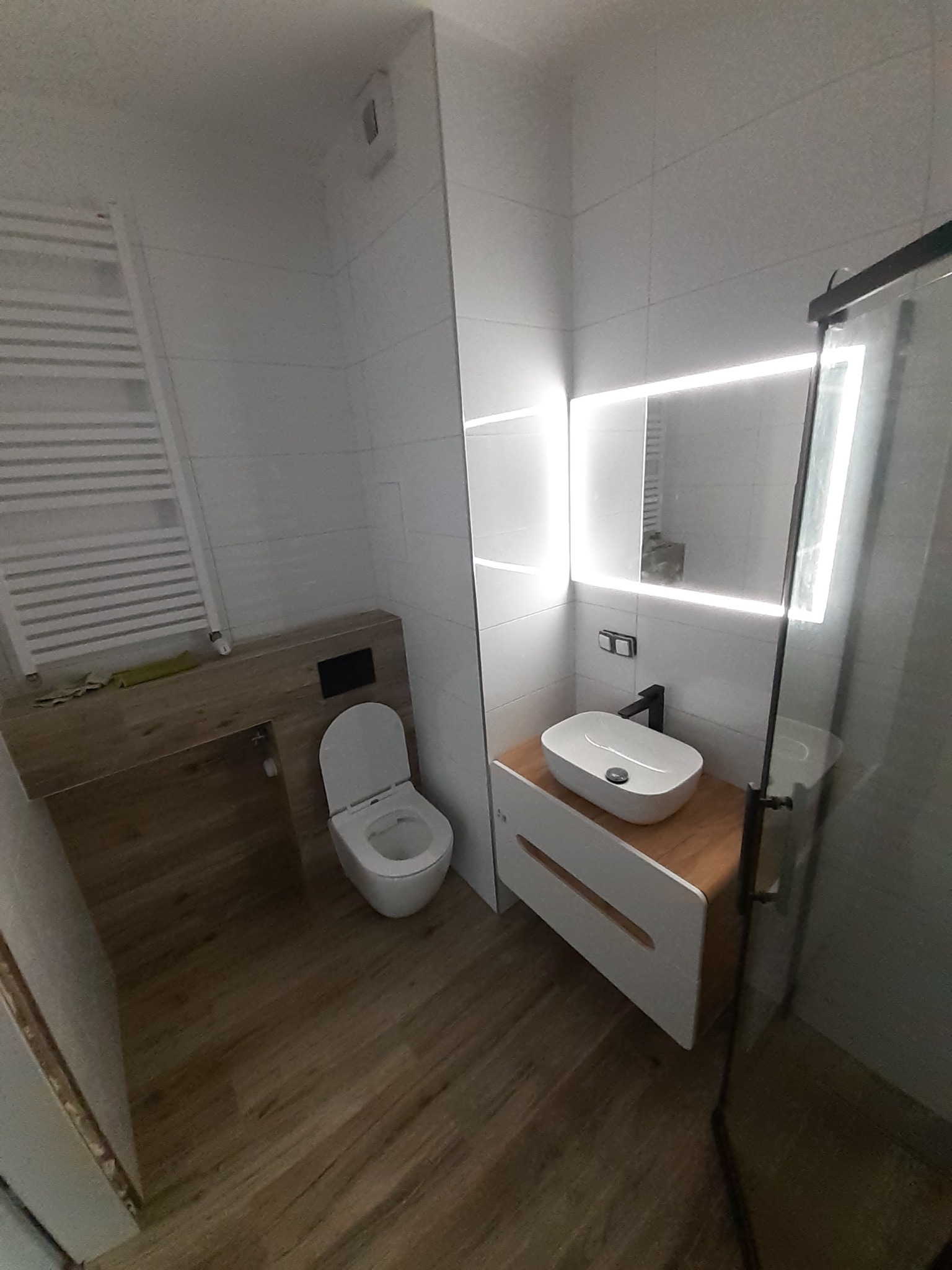 Nowoczesna łazienka z podświetlanym lustrem, umywalką nablatową i toaletą w zabudowie. Jasne płytki i drewnopodobna podłoga tworzą spójną całość.