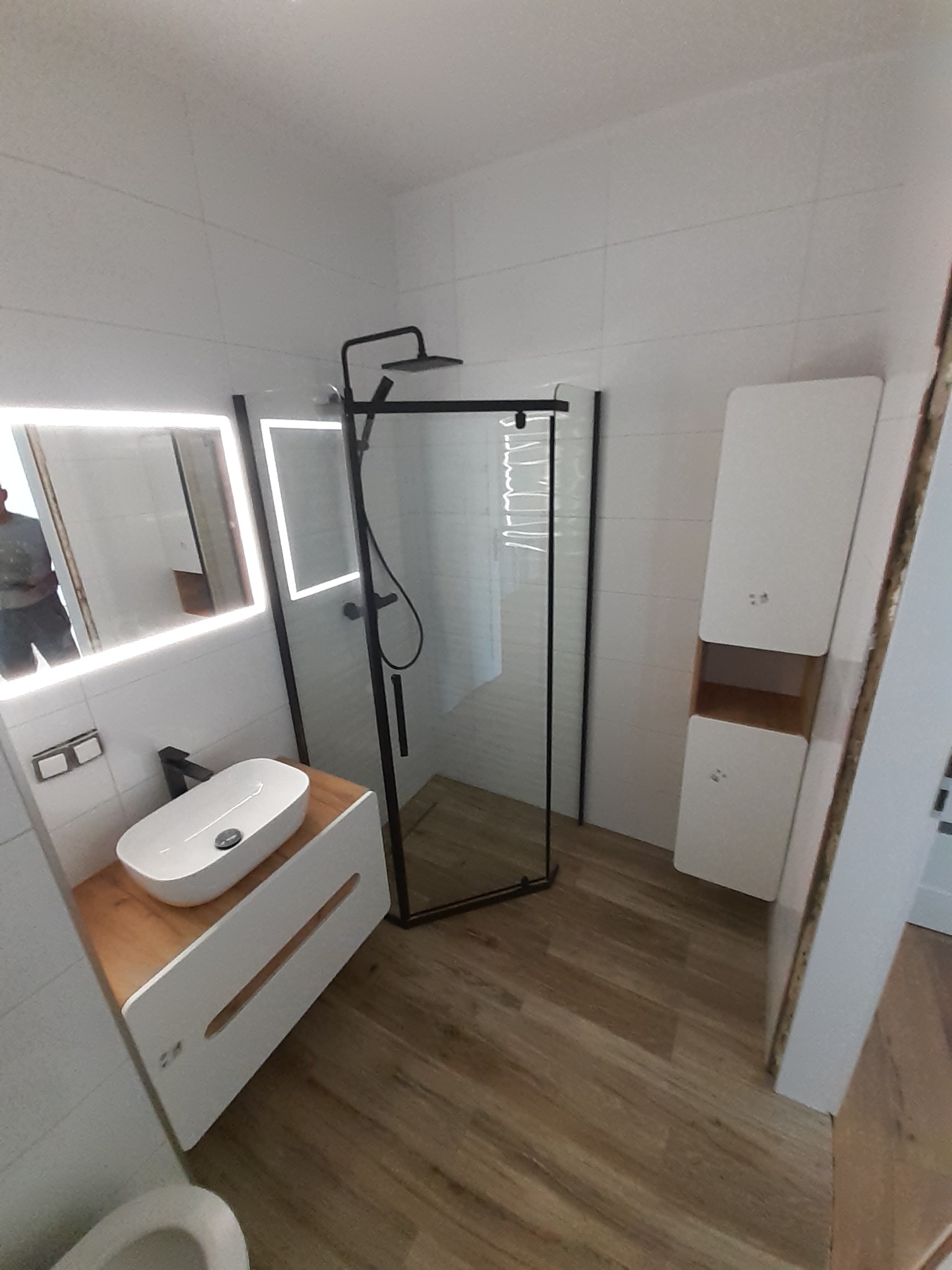 Nowoczesna łazienka z kabiną prysznicową typu walk-in, umywalką nablatową na drewnianym blacie i podświetlanym lustrem. Białe płytki i drewnopodobna podłoga.