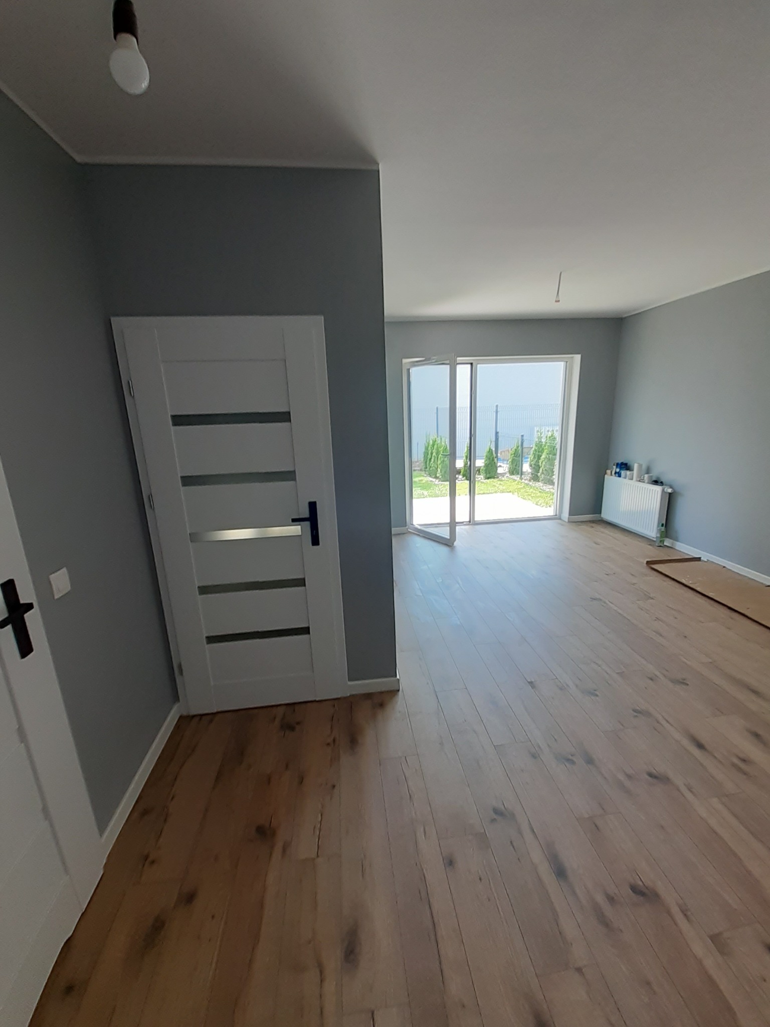 Jasne drewniane panele podłogowe w szarym pokoju z białymi drzwiami i widokiem na ogród przez otwarte drzwi balkonowe. Minimalistyczny design.