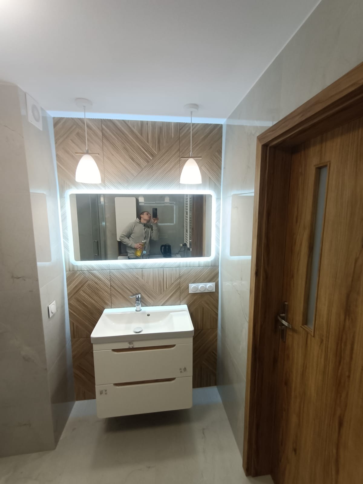 Nowoczesna łazienka po remoncie: biała szafka z umywalką, podświetlane lustro, drewniana mozaika na ścianie i drewniane drzwi. Widoczne oświetlenie.