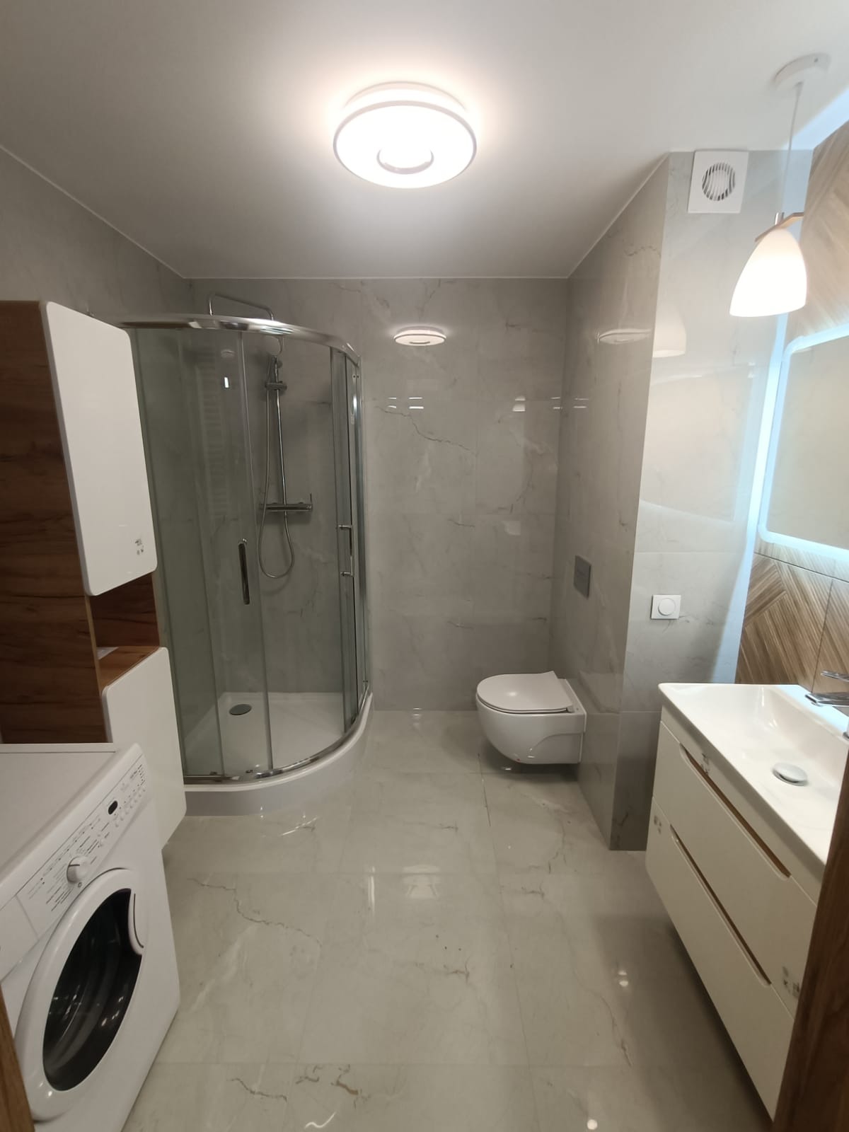 Wyremontowana łazienka w Bydgoszczy z kabiną prysznicową, umywalką z szafką i toaletą w odcieniach szarości i bieli. Widoczna pralka i oświetlenie sufitowe.