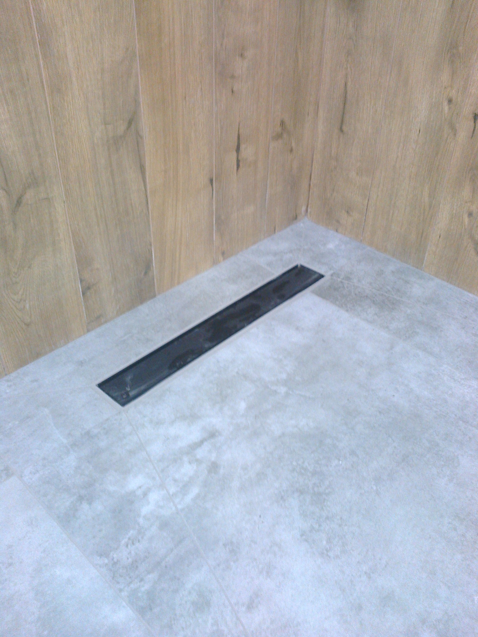 Nowoczesna łazienka z szarymi płytkami i odpływem liniowym. Ściana wyłożona panelami imitującymi drewno. Minimalistyczny design, jasne wnętrze.