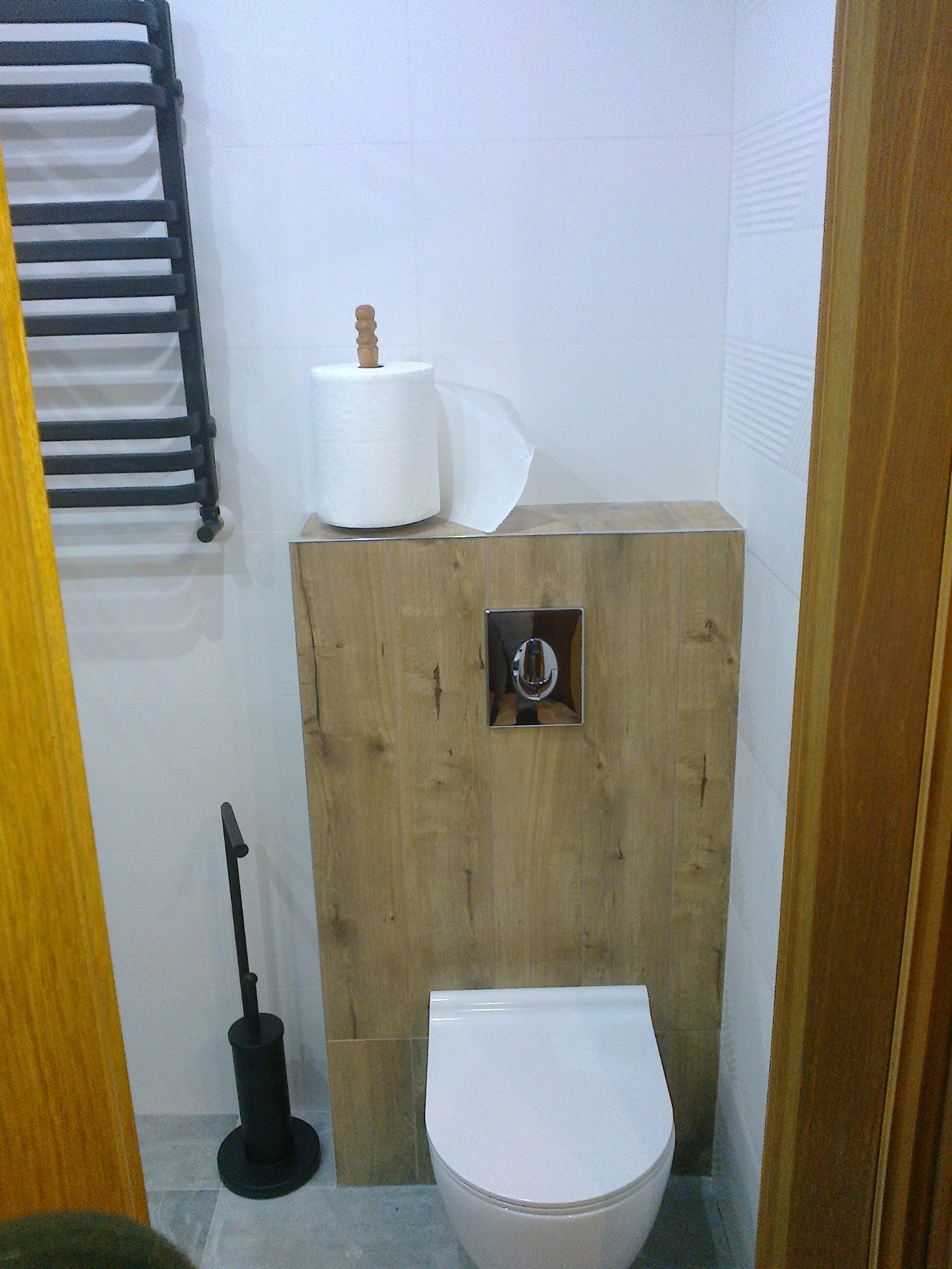 Nowoczesna toaleta podwieszana z drewnianą obudową, chromowanym przyciskiem i grzejnikiem drabinkowym w wykończonej łazience z jasnymi płytkami i akcesoriami.