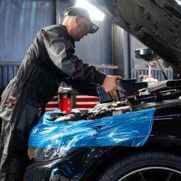 SEBASTIAN WACHECKI PowerUP Mechanika Pojazdowa - Czarny samochód z otwartą maską, zabezpieczony folią. Mechanik w kombinezonie dolewa płyn eksploatacyjny. Warsztat z czerwonymi elementami w tle.