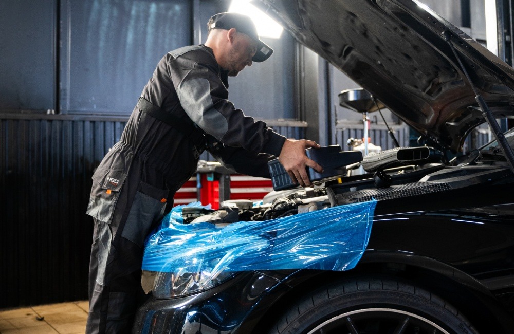 Czarny samochód z otwartą maską, zabezpieczony folią. Mechanik w kombinezonie dolewa płyn eksploatacyjny. Warsztat z czerwonymi elementami w tle.