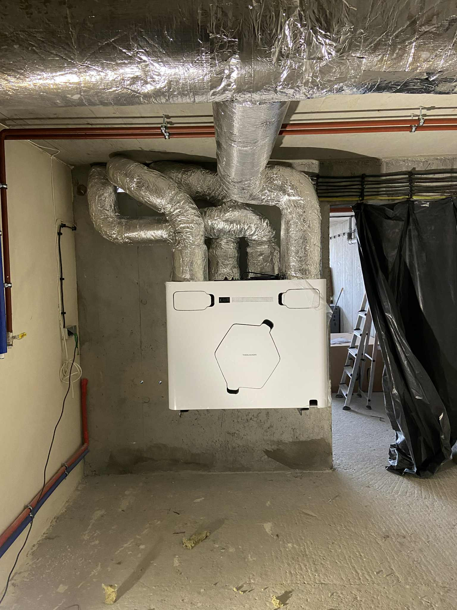 Biała centrala wentylacyjna z podłączonymi, izolowanymi przewodami w surowym pomieszczeniu. Widoczne rury i kable na ścianach, drabina w tle.