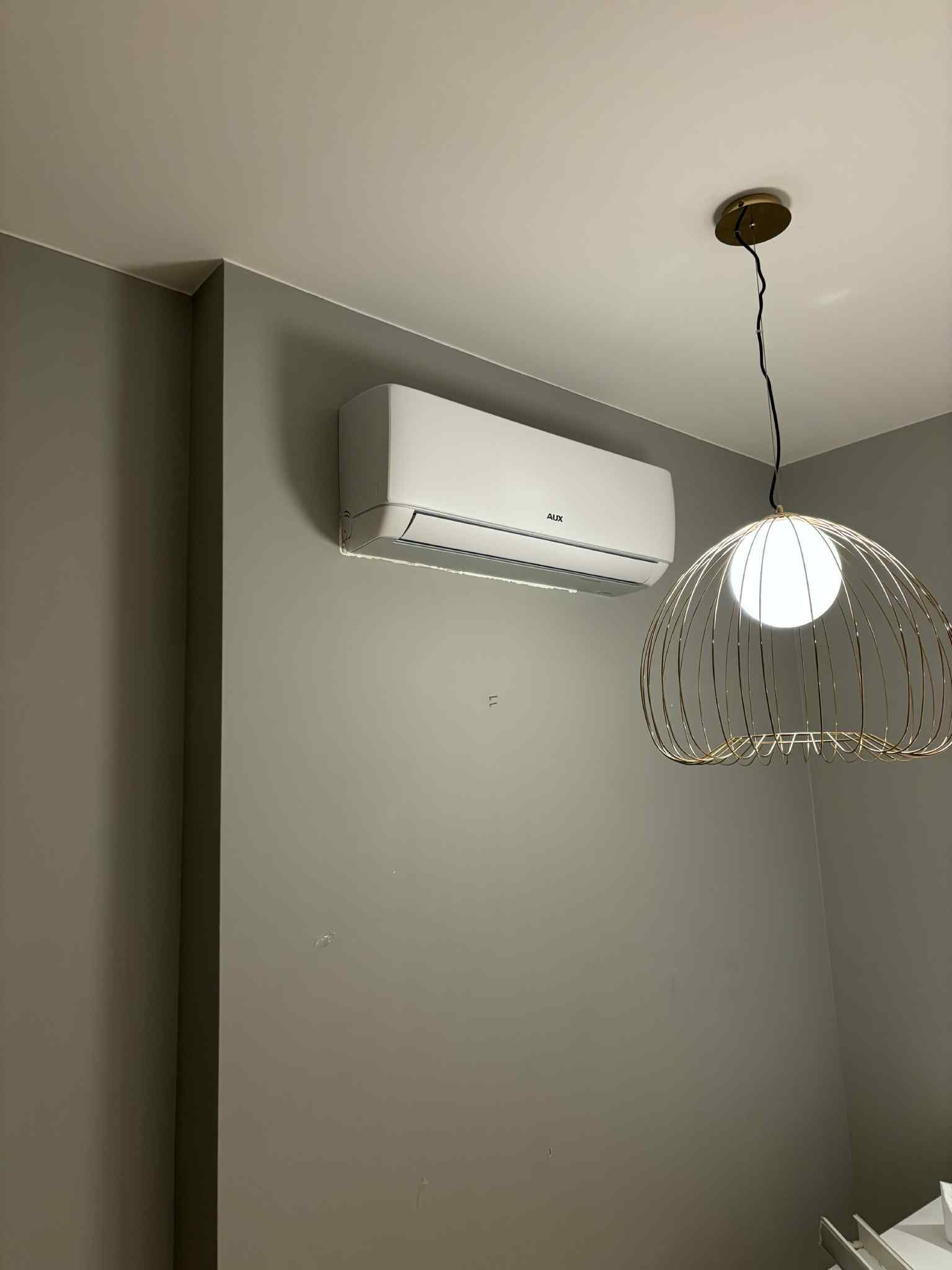 Biały klimatyzator AUX na szarej ścianie w pokoju, obok designerska lampa z metalowym kloszem. Widok z dołu, oświetlenie dzienne.