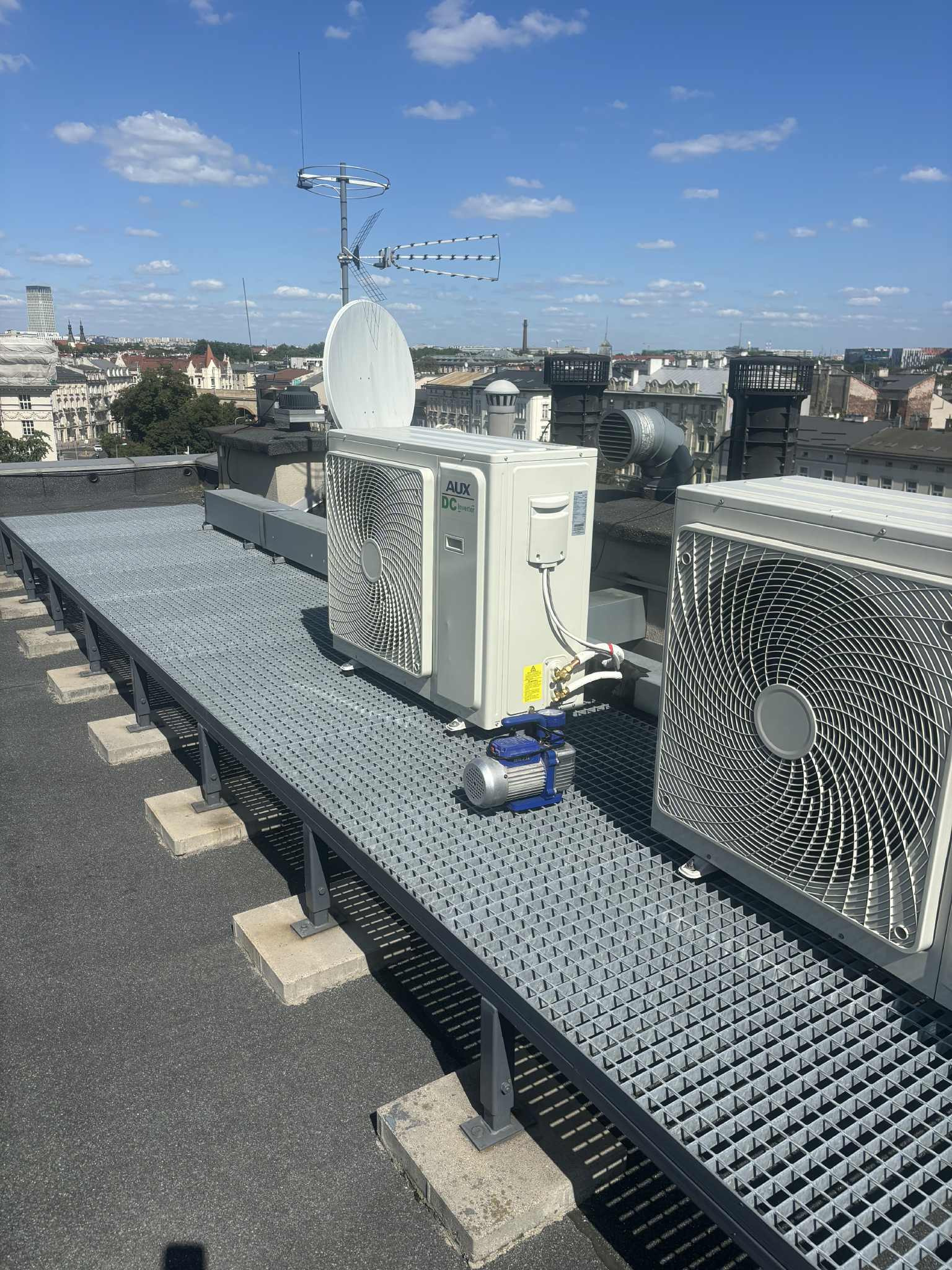 Białe jednostki zewnętrzne klimatyzacji AUX DC Inverter na dachu budynku, z widoczną pompą próżniową i anteną satelitarną w tle, w słoneczny dzień.