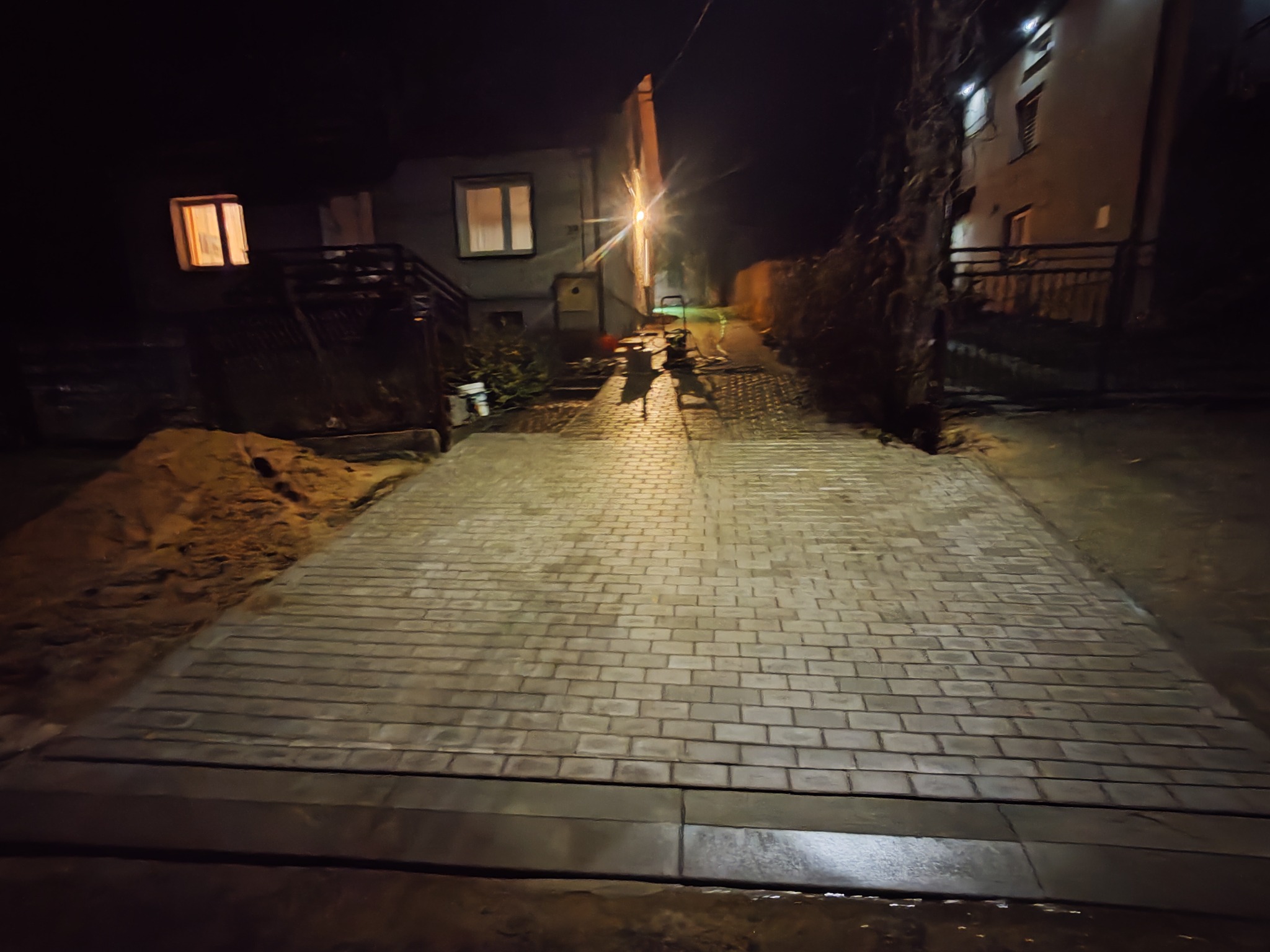 Ułożona kostka brukowa w ciemności, oświetlona lampą uliczną, prowadząca wzdłuż domu. Widoczny stos piasku i narzędzia brukarskie w tle.