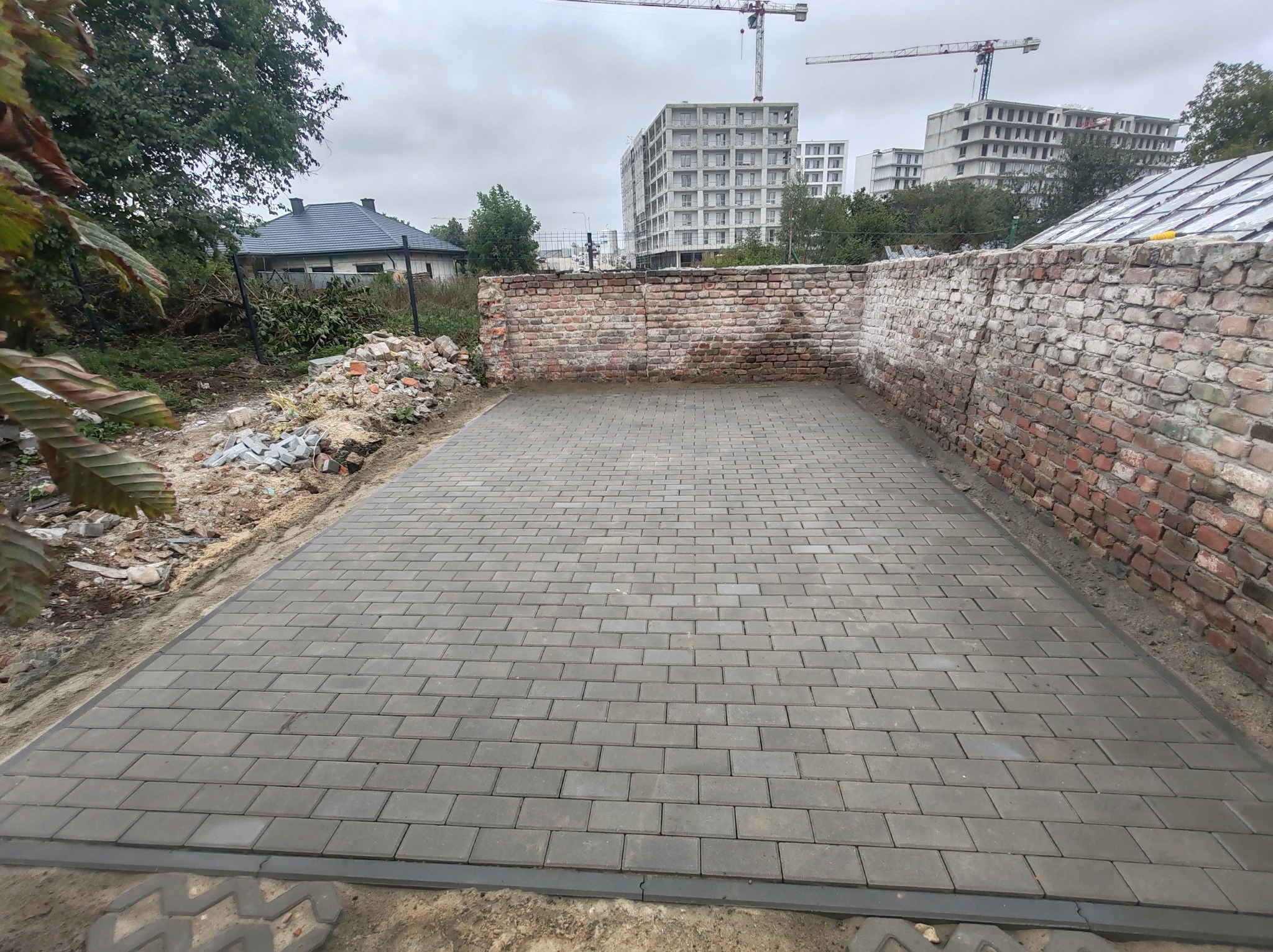 Ułożona szara kostka brukowa na posesji z ceglanym murem w tle, widok z góry. Na drugim planie budynki w budowie i żurawie.