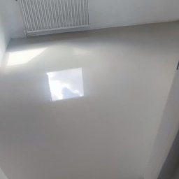 Rafał Muller LOGOS - Jasnoszara, gładka wylewka samopoziomująca w pokoju z oknem dachowym i grzejnikiem. Widoczne przygotowania do dalszych prac wykończeniowych podłogi.