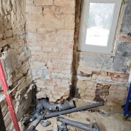 Rafał Muller LOGOS - Remont łazienki w trakcie: odsłonięta cegła, poziomica, stelaż podtynkowy WC i brodzik. Widoczne rury kanalizacyjne. Surowy stan, przygotowanie do dalszych prac.