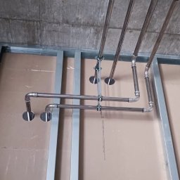 Rafał Muller LOGOS - Metalowa konstrukcja sufitu podwieszanego z instalacją rur, widoczne przewody elektryczne i płyty kartonowo-gipsowe. Ujęcie z dołu, surowy stan wykończenia.
