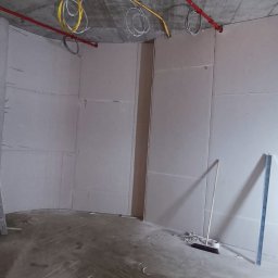 Rafał Muller LOGOS - Wnętrze w trakcie prac z płytami gipsowo-kartonowymi. Widoczna drabina, profile metalowe i częściowo zaokrąglona ściana. Instalacje pod sufitem.