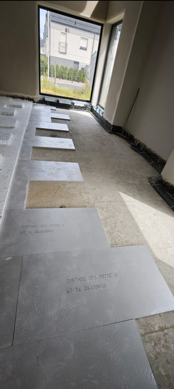 Przygotowanie podłoża pod panele: rozłożone płyty izolacyjne SYNTHOS XPS PRIME B w surowym wnętrzu z dużym oknem. Widoczne kable i elementy instalacji przy ścianie.