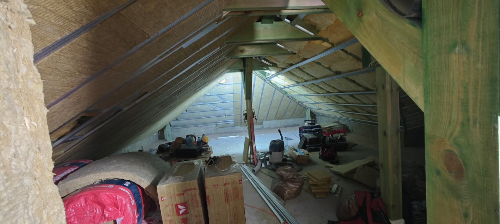 Izolacja poddasza wełną mineralną, widoczne krokwie i metalowe profile. Częściowo ułożona izolacja, narzędzia i materiały budowlane na podłodze.