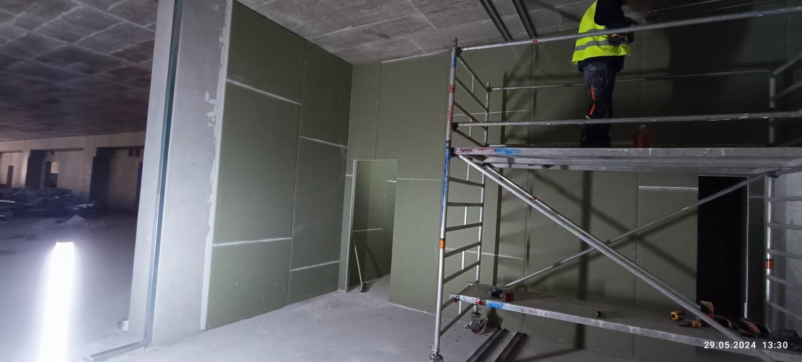Montaż płyt karton gips na ścianach w surowym wnętrzu. Widoczne rusztowanie i pracownik w odblaskowej kamizelce. Częściowo wykończone ściany z płyt G-K.