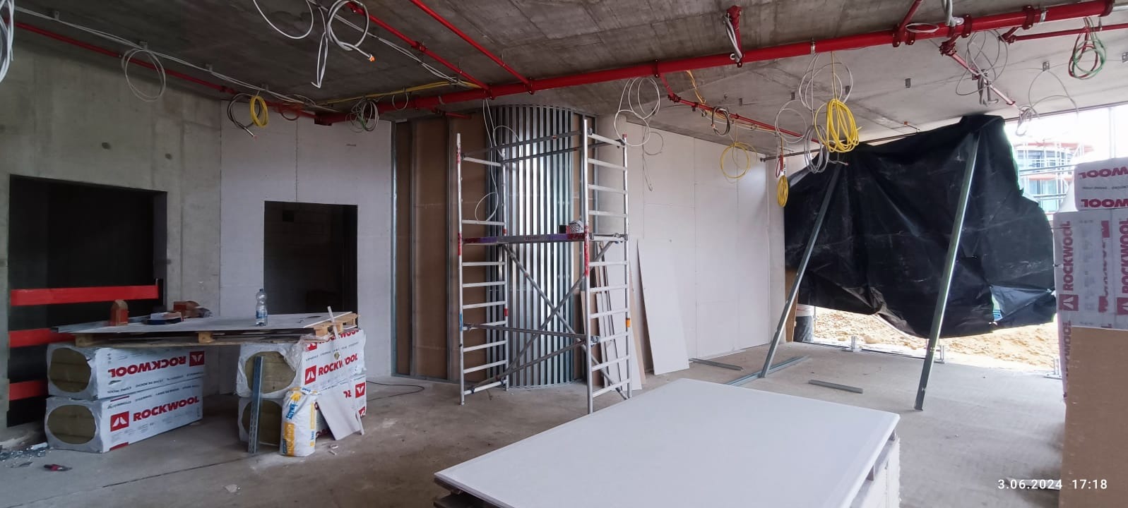 Wnętrze w trakcie prac wykończeniowych: widoczne płyty karton-gips, rusztowanie, instalacje pod sufitem i materiały izolacyjne Rockwool. Surowy beton i okna w tle.
