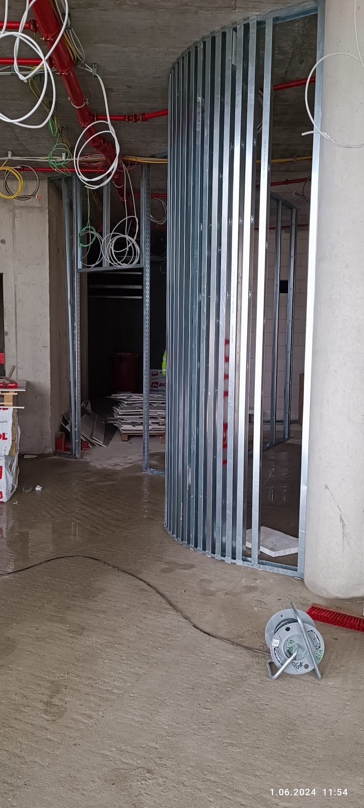 Metalowa konstrukcja ścianki działowej na mokrej posadzce, widoczne instalacje pod sufitem i materiały budowlane w tle. Etap prac wykończeniowych w surowym wnętrzu.