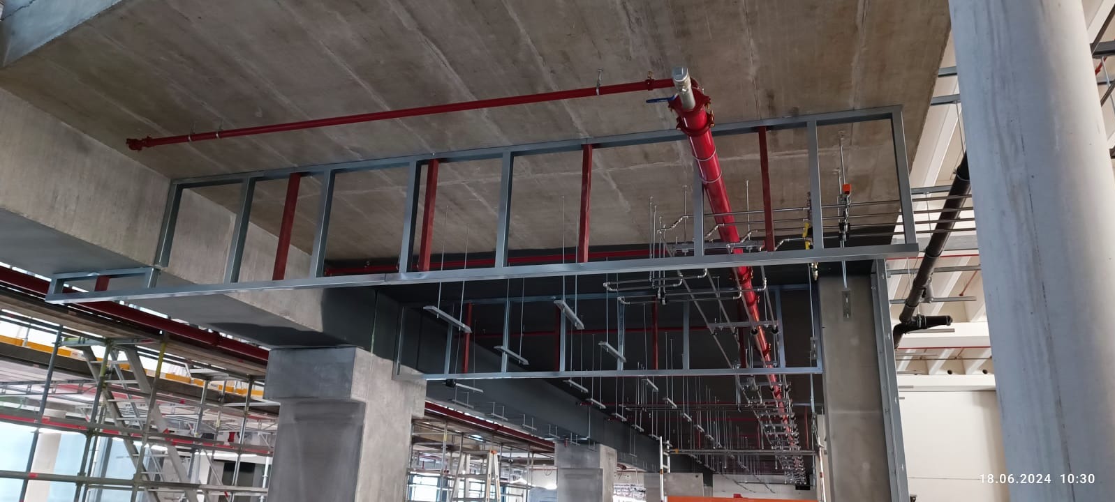 Sufit w trakcie montażu instalacji: czerwone rury, metalowa konstrukcja podwieszana, betonowe filary. Widoczne elementy rusztowania i inne instalacje.