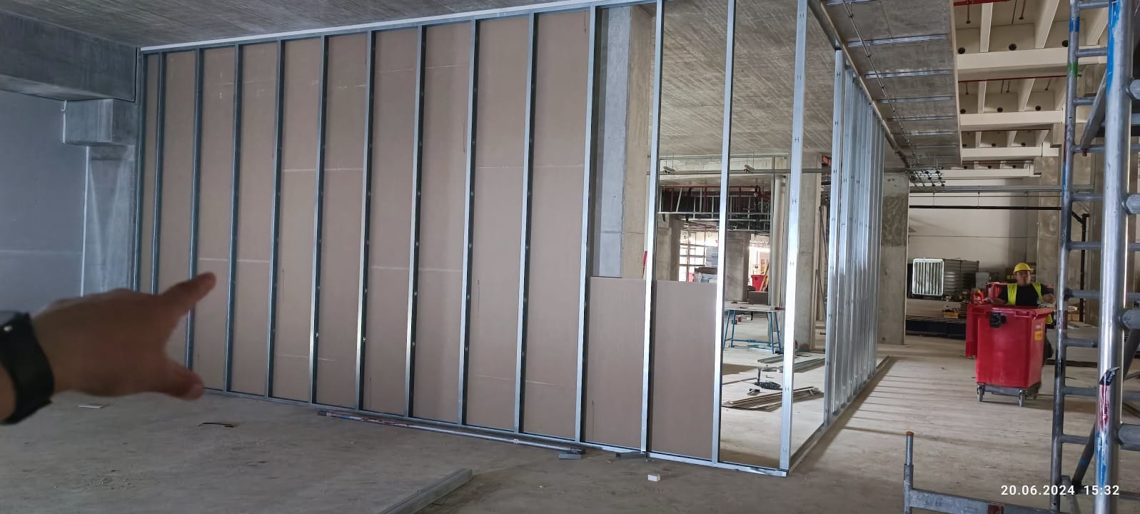 Montaż ścian działowych z płyt gipsowo-kartonowych na metalowym stelażu w surowym wnętrzu. Widoczny fragment dłoni wskazującej na konstrukcję.