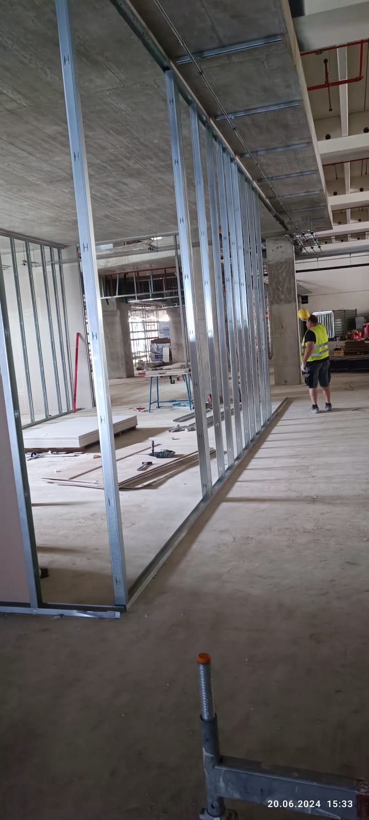 Montaż metalowej konstrukcji ścian działowych w surowym wnętrzu. Widoczny pracownik w oddali. Elementy konstrukcji z profili stalowych, surowy betonowy strop.