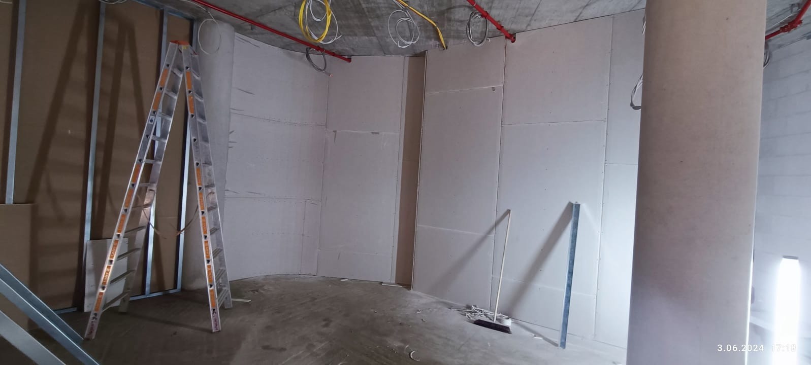 Wnętrze w trakcie prac z płytami gipsowo-kartonowymi. Widoczna drabina, profile metalowe i częściowo zaokrąglona ściana. Instalacje pod sufitem.