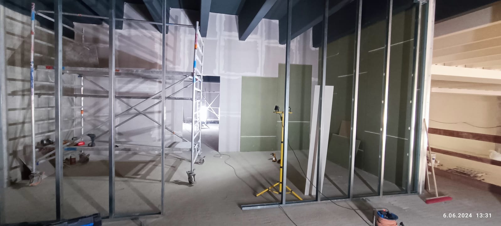 Wnętrze w trakcie prac wykończeniowych: metalowa konstrukcja, częściowo zamontowane płyty gipsowo-kartonowe, rusztowanie. Widoczne narzędzia i oświetlenie budowlane.